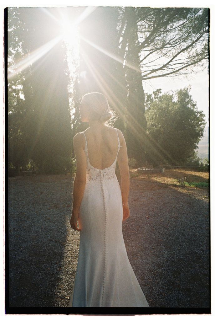 A bride in a backless white wedding dress stands outdoors, illuminated by sunlight streaming through trees.